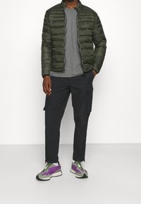 Grön puffjacka med vadderad textur, grå T-shirt, svarta cargobyxor med fickor och flerfärgade sneakers med lila detaljer.