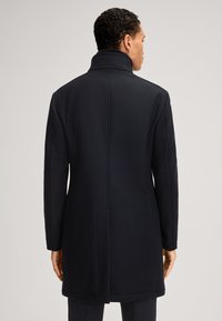 Cappotto in lana navy con colletto alto, fit su misura e texture liscia. Presenta linee pulite e uno stile minimalista, senza bottoni visibili.