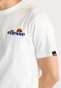 T-shirt bianca in cotone con logo "ellesse" ricamato in blu nautico e rosso; maniche corte con etichetta nera brandizzata sul polsino.