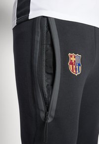Zwarte sweatpants met ritssluitingen aan de zijzakken, gemaakt van een gestructureerde stof en voorzien van een kleurrijk logo van de voetbalclub Barcelona.