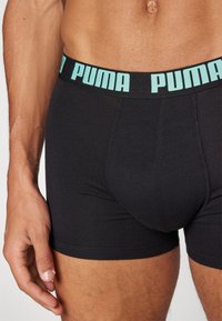 Black cotton trunk with a light blue "PUMA" logo waistband. Features a fitted design and smooth texture. Stitching is visible along edges.