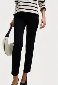 Pantalon noir à coupe slim, avec une texture de tissu lisse. Porté avec un haut à manches longues rayé et un sac à main blanc.