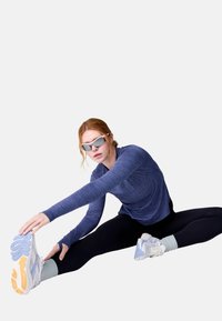 Sportinė apranga su prigulusia mėlyna ilgomis rankovėmis palaidine, juodomis leggingomis ir baltais bateliais su violetiniais akcentais. Modelis atlieka tempimo pratimus.