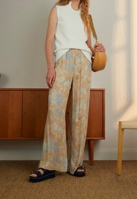 Person med ärmlös vit stickad topp, vida byxor med blommönster, svarta plattformssandaler och håller en tanaxelväska inomhus.