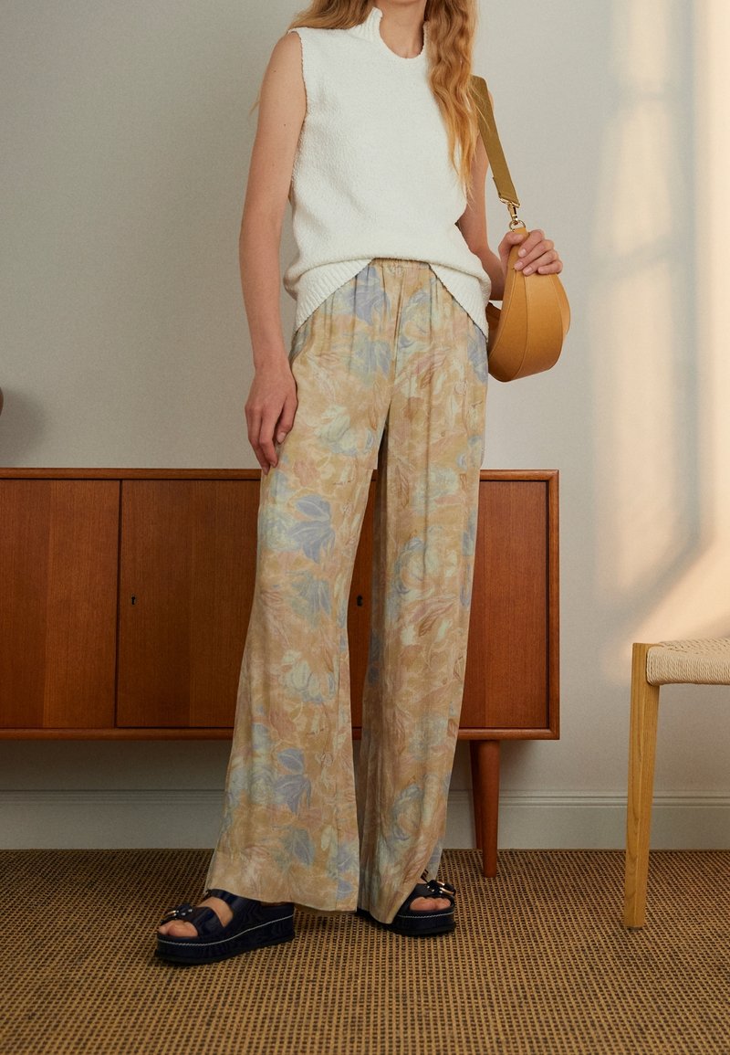 Person med ärmlös vit stickad topp, vida byxor med blommönster, svarta plattformssandaler och håller en tanaxelväska inomhus.