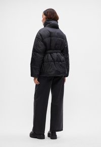 Casaco puffer preto com gola alta e cintura cingida, apresentando secções acolchoadas. Combinado com jeans largos de cor escura e sapatos pretos robustos.