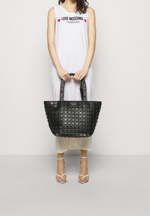 Nainen valkoisessa hihattomassa mekossa, jossa on "Love Moschino" -teksti, pitelee suurta mustaa pehmeästi tikattua olkalaukkua, jossa on merkkikahvat. Valkoiset korkokengät.