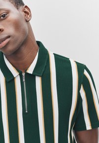 Grün-weiß gestreiftes kurzärmeliges Poloshirt mit goldenen Akzenten, ausgestattet mit einem Reißverschluss und einer gerippten Textur im Stoff.