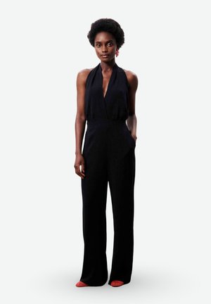 Femme aux cheveux courts et bouclés portant une combinaison noire sans manches à col halter et des chaussures rouges à bout pointu, debout devant un fond blanc.