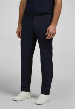 Mann trägt eng anliegende dunkelblaue Hose und weiße Turnschuhe, steht vor einem schlichten hellgrauen Hintergrund.