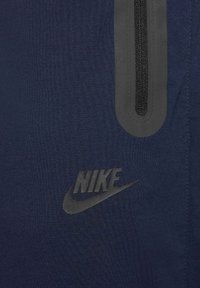 Granatowa tkanina o gładkiej fakturze z kieszenią na zamek oraz czarnym logo Nike w dolnym rogu. Subtelne oznakowanie i minimalistyczny design.