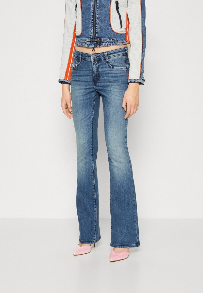 Personne portant un jean bleu taille mi-haute, coupe droite avec décoloration, une veste courte bleu et blanc avec des rayures orange, et des talons pointus roses.