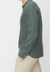 Lniana koszula oliwkowa zielona z zapiętym na guziki kołnierzykiem, długimi rękawami, kieszenią na piersi i detalami przy mankietach. Charakteryzuje się luźnym krojem i gładką fakturą.