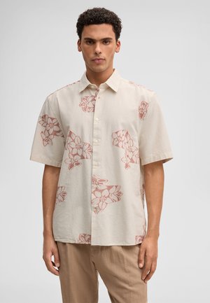 Mann trägt ein beige kurzärmliges Hemd mit roten Blumenmustern und hellbraune Hose, steht vor einem einfarbigen Hintergrund.