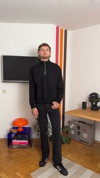 Sweatshirt preto com fecho, colarinho alto, combinado com calças pretas e sapatos pretos com riscas brancas. Design minimalista, sem padrões visíveis.