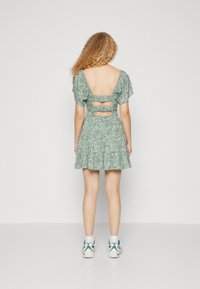 Osoba s kudrnatými vlasy stojí čelem k nám, má na sobě zelené květinové šaty s nafouknutými rukávy a výstřihem na zádech, doplněné bílými teniskami.