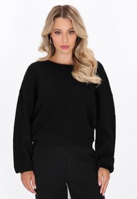 Schwarzer Pullover aus dickem, strukturiertem Material mit Boot-Ausschnitt, überschnittenen Schultern und lockerer Passform. Kombiniert mit dunklen Hosen.