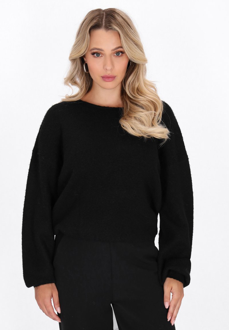 Schwarzer Pullover aus dickem, strukturiertem Material mit Boot-Ausschnitt, überschnittenen Schultern und lockerer Passform. Kombiniert mit dunklen Hosen.