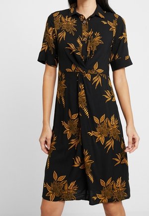 Zwarte knielange jurk met grote gele bloemenprint, korte mouwen, kraag, knopen en een gedraaide knoop vooraan in de taille.
