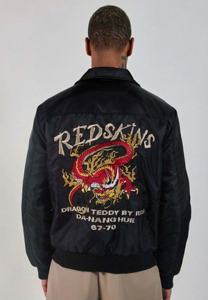 Homme portant une veste noire avec un dragon brodé en rouge et or et le texte "REDSKINS" au dos, debout devant un fond uni.