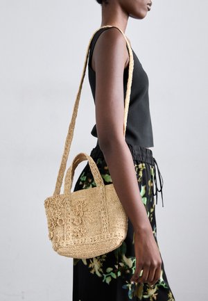 Small beige woven bag with floral crochet details and dual handles, carried on the shoulder. The model wears a black top and floral skirt.