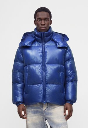 Ung man med kort lockigt hår iklädd en glänsande blå vadderad jacka och lätt tvättade jeans, stående mot en enkel ljus bakgrund.