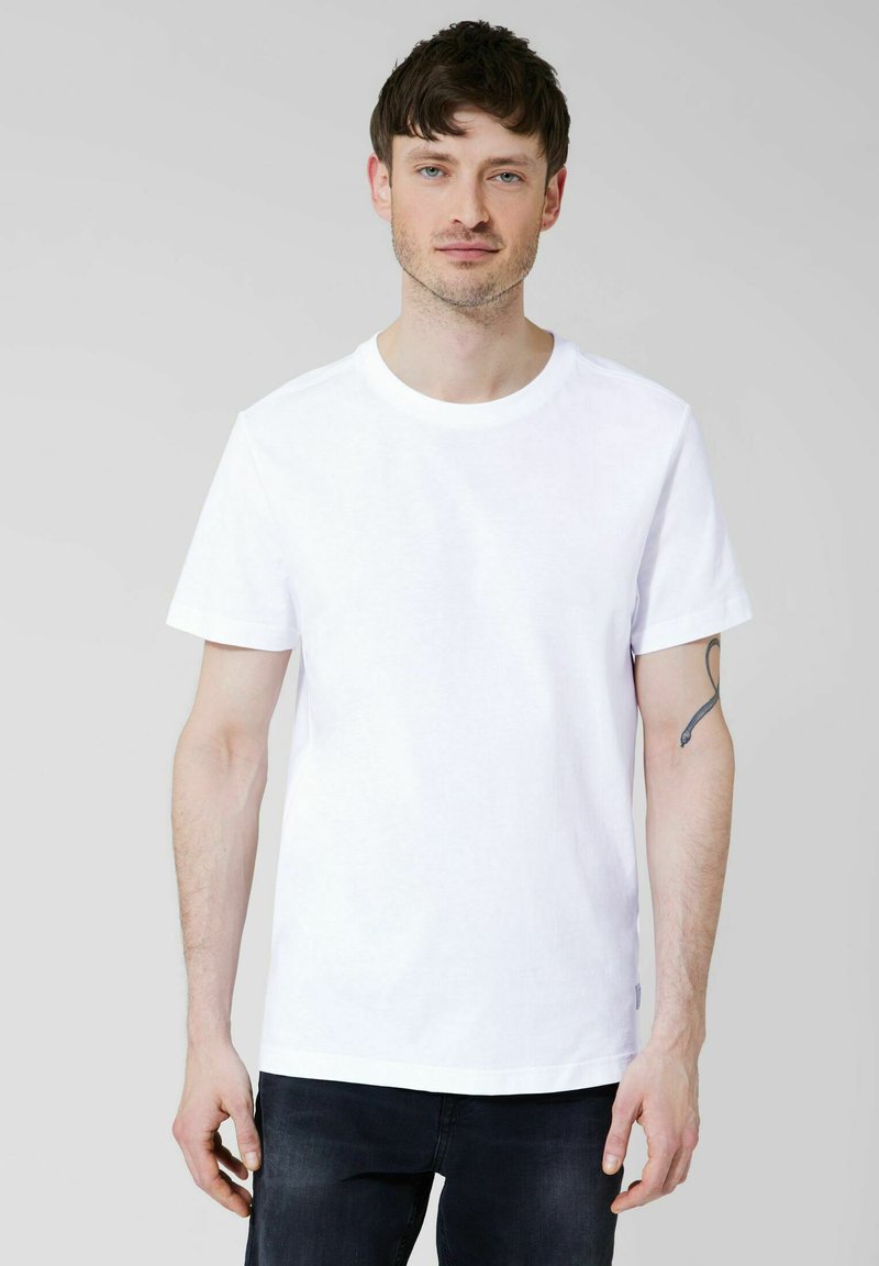Hombre con cabello castaño corto que lleva una camiseta blanca de manga corta y jeans negros, posando contra un fondo gris claro.