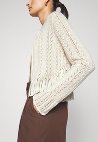 Knit cardigan in cream with a textured, open weave pattern, featuring long sleeves and a relaxed fit. Paired with brown trousers.