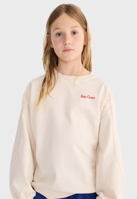 Joven niña con cabello rubio largo viste una sudadera crema con el logo rojo "Bobo Choses", de pie frente a un fondo liso de color gris claro.