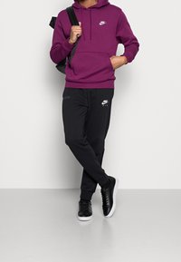 Persona vestida con una sudadera morada de Nike, pantalones de chándal negros de Nike, zapatillas negras y llevando una mochila negra sobre un hombro.