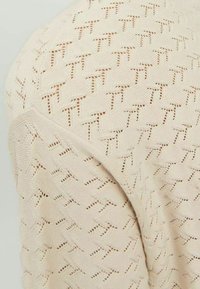 Beige gebreid kledingstuk met een open weefpatroon, diagonale lijnen en kleine uitsnijdingen. Zachte textuur, lichtgewicht en casual ontwerp.