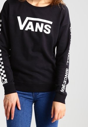 Personne portant un sweat-shirt Vans noir avec un logo blanc sur la poitrine et un texte sur les manches, associé à un jean bleu, sur un fond uni.