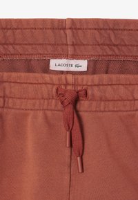 Pantalons joggers en terre cuite avec un tissu texturé, taille élastique et cordon de serrage avec embouts en plastique ; étiquette logo Lacoste cousue en haut.