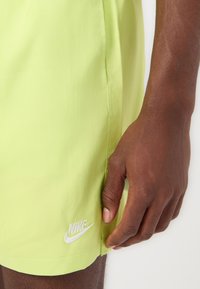 Neonvihreät urheilushortsit, joissa on sileä pinta, sivuvetoketju ja valkoinen brodeerattu Nike-logo alhaalla jalassa.