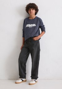 Ciemnoszara koszulka z długim rękawem z napisem "ŚWIATOWA SPOŁECZNOŚĆ" w białym kolorze, zestawiona z czarnymi luźnymi dżinsami i białymi trampkami z czarnymi akcentami.