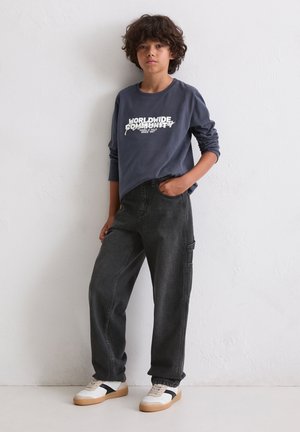 Dunkelgraues langärmeliges Shirt mit dem Aufdruck "WELTWEITE GEMEINSCHAFT" in Weiß, kombiniert mit schwarzen baggy Jeans und weißen Sneakers mit schwarzen Akzenten.