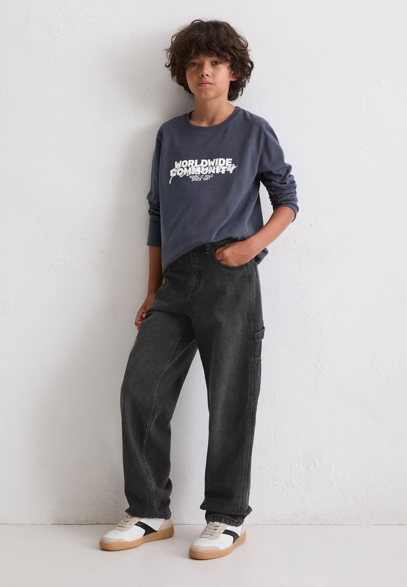 Ciemnoszara koszulka z długim rękawem z napisem "ŚWIATOWA SPOŁECZNOŚĆ" w białym kolorze, zestawiona z czarnymi luźnymi dżinsami i białymi trampkami z czarnymi akcentami.