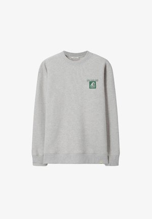 Jeune homme portant un sweat-shirt beige, une chemise à rayures vertes, un jean bleu retroussé et des chaussures marron, debout, une main dans la poche, l'autre touchant ses cheveux.