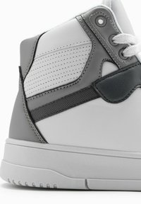 Sneaker montante en cuir blanc lisse avec des accents gris, panneaux texturés, oeillets renforcés et semelle en caoutchouc avec des rainures horizontales.