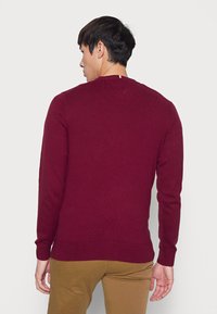 Bordeauxfarbener Strickpullover mit langen Ärmeln und Rundhalsausschnitt. Verfügt über eine V-förmige Naht auf der Rückseite und einen dezenten farbigen Streifen am Hals.