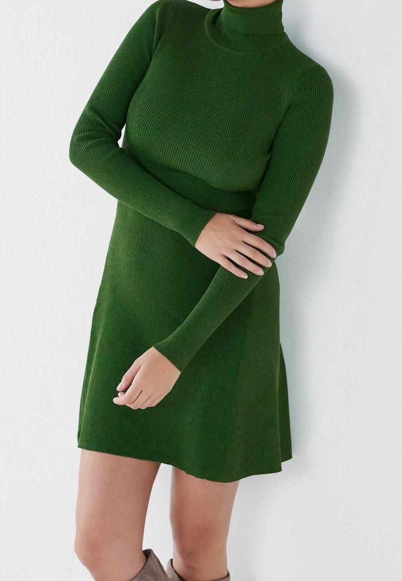 Robe tricot à côtes verte avec un col montant et des manches longues, présentant un corsage ajusté et une jupe évasée. Texture douce et coupe décontractée.