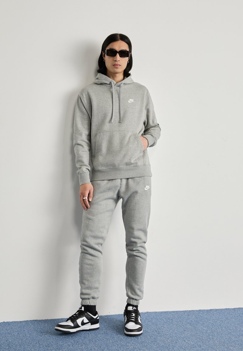 Grau Sweatshirt und Sweatpants, beide mit einem kleinen weißen Nike-Logo. Getragen mit schwarzen Sneakers mit weißen Akzenten. Glatte Textur.