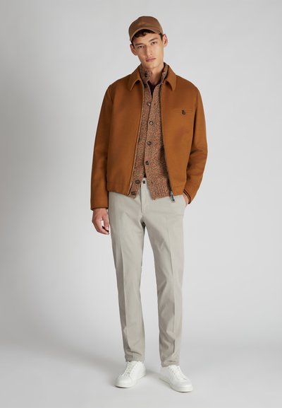 Brązowa kurtka z wysokim kołnierzem, beżowa koszula w wzory, jasnoszare spodnie i białe trampki; gładka faktura, swobodny fason.