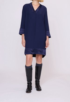 Femme portant une robe bleu marine longueur genou avec un bord en satin, associée à des bottes noires montantes et des chaussettes sombres.