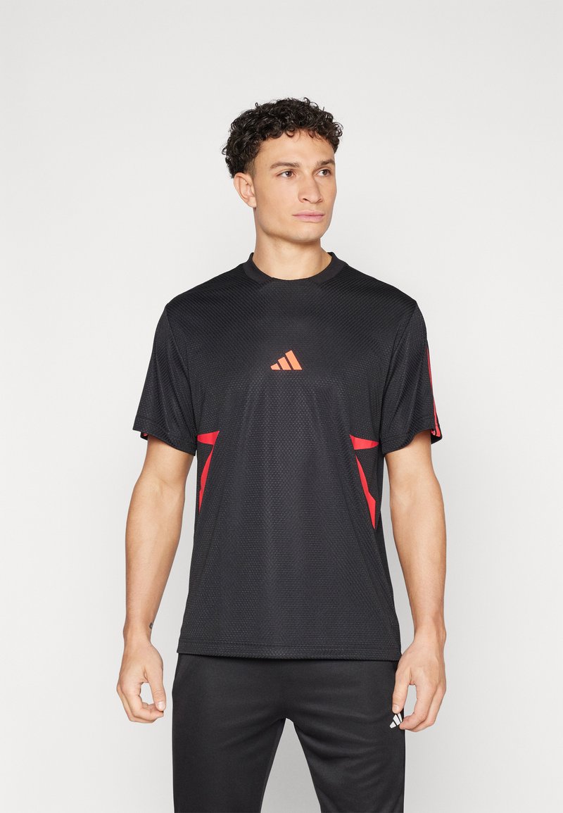 Čierne športové tričko s textúrou sieťoviny, s červenými akcentmi a oranžovým logom Adidas na hrudi. Spárované s čiernymi nohavicami.