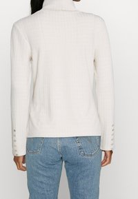 Top blanco de cuello alto con mangas largas, que presenta botones decorativos en los puños. Combinado con jeans de color azul claro.