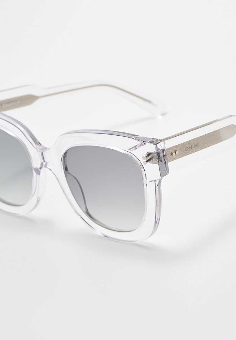 Gafas de sol transparentes de gran tamaño con lentes grises degradados, montura gruesa y acentos metálicos. Presentan el nombre de la marca grabado en la patilla.