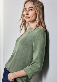 Světlé zelené pletené tričko s texturovaným povrchem, 3/4 rukávy a kulatým výstřihem. Spárováno s tmavě modrými džíny, nošeno neformálně.