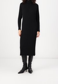 Robe en tricot côtelé noire avec un col montant et des manches longues, dotée d'une fente latérale. Associée à des bottines noires à talons pointues.