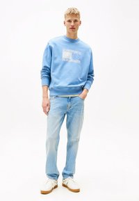 Sudadera azul claro con un diseño gráfico texturizado en el pecho, combinada con jeans de lavado claro y zapatillas blancas con suela marrón.